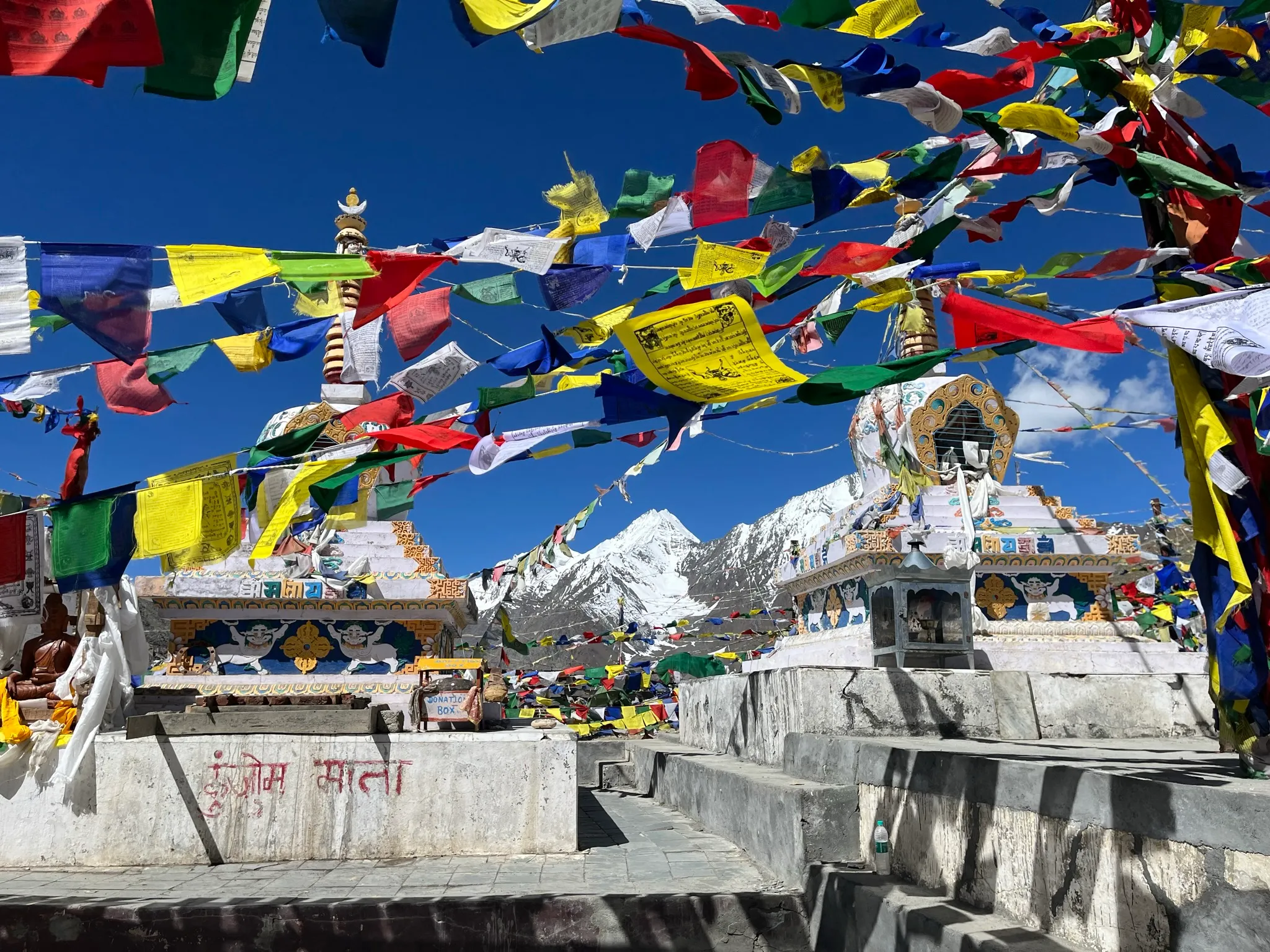Whitewashed Tibetan Buddhist stupas strung with hundreds of colorful prayer flags, framing snow-capped Himalayan peaks under a deep blue sky in Ladakh